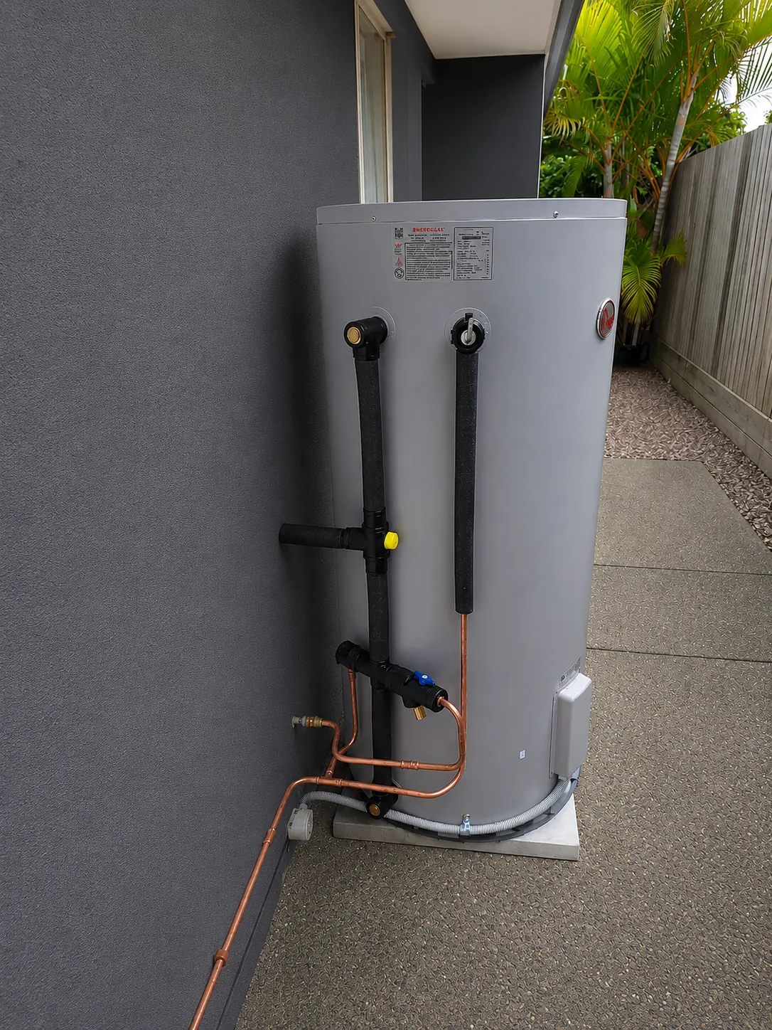 Rheem electric storage hot water unit installed against a rendered wall beside a path